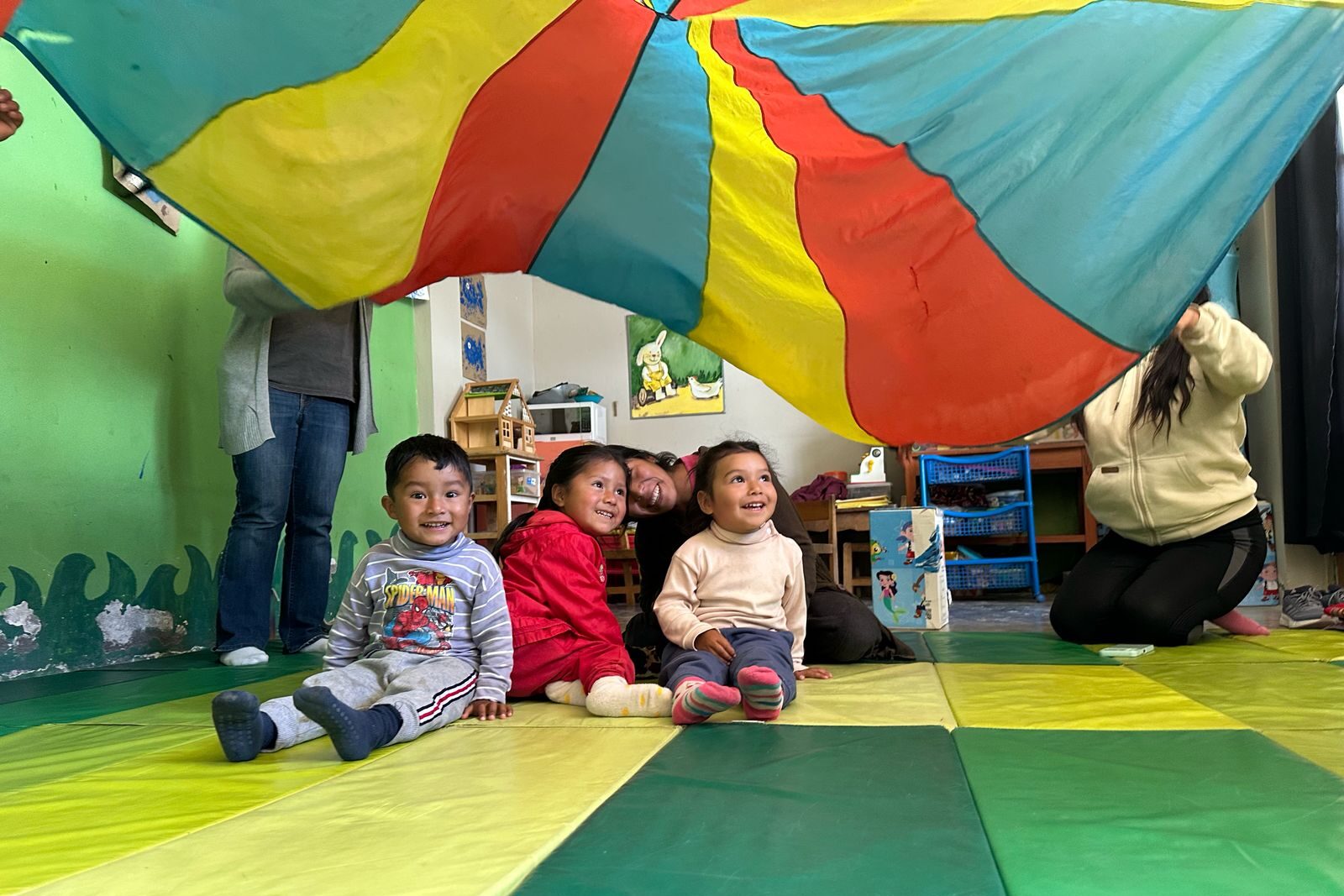 Drie kleuters zijn samen met hun mama's aan het spelen. Er wordt boven de hoofden van de kinderen een doek gewapperd. Iedereen is aan het lachen.